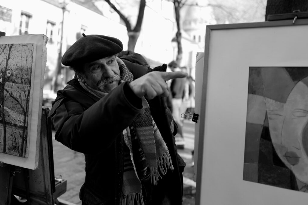 Montmartre. Photo en noir et blanc d'un homme avec un berret montrant du doigt une œuvre. 